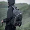 A person wearing a black jacket and pants is carrying a black waterproof hiking backpack with a laptop sleeve in an outdoor, 