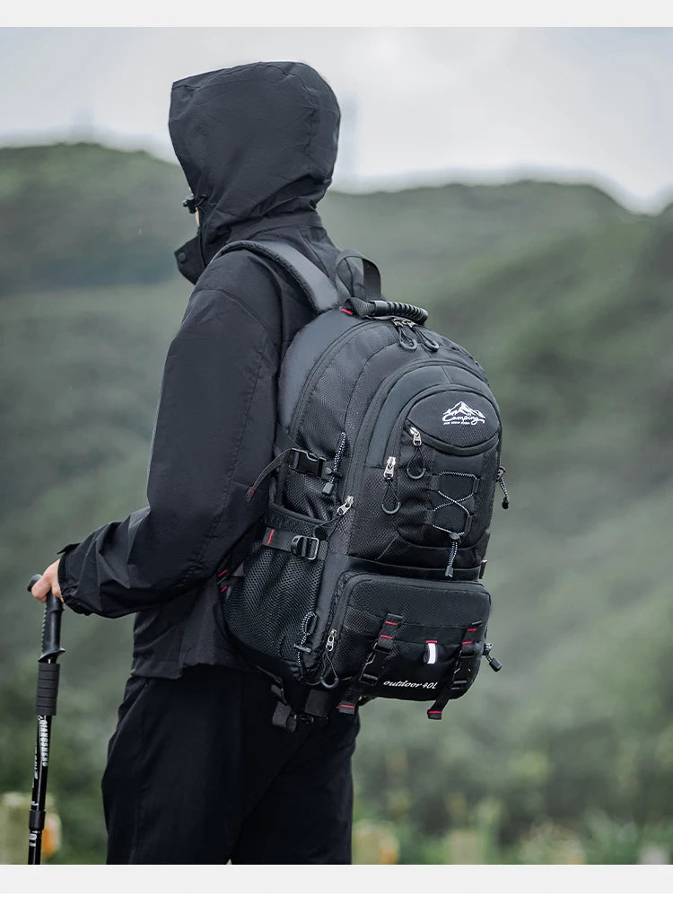 A person wearing a black jacket and pants is carrying a black waterproof hiking backpack with a laptop sleeve in an outdoor, 
