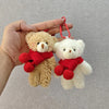 A hand holds a fluffy brown teddy bear and a white teddy bear, both with red scarves, as a keychain.