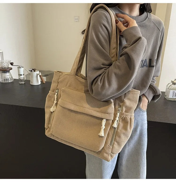 A person is holding a tan corduroy shoulder bag with a zipper on their shoulder while standing in a kitchen setting.