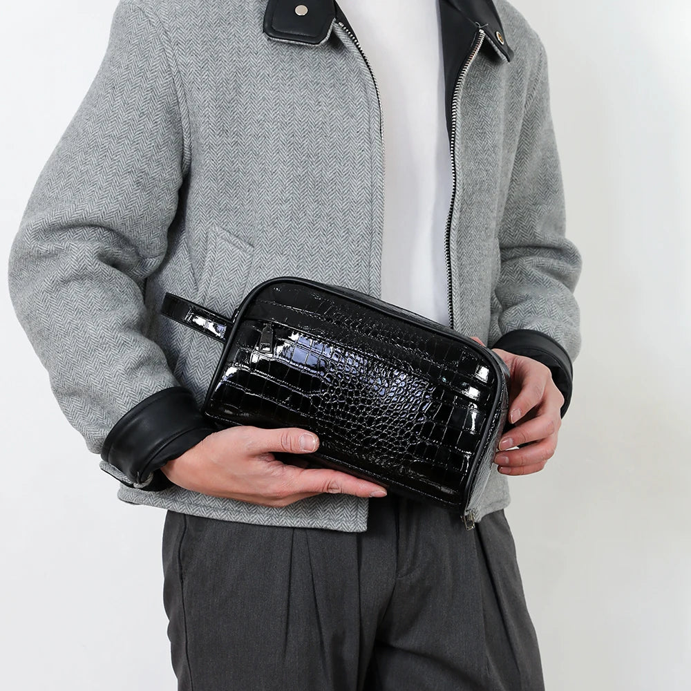 A person is holding a black crocodile print PU leather clutch bag against a gray blazer and dark pants.