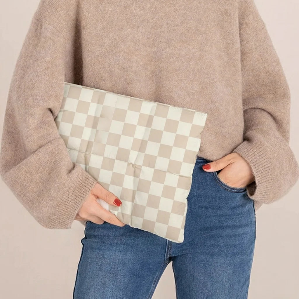 Une personne portant un pull beige tient une housse d'ordinateur à carreaux contre son jean.