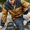 A male model wearing a tan and black tactical jacket and dark pants, holding a gray water bottle outdoors.
