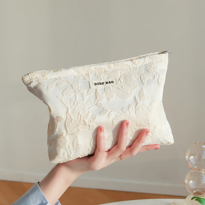 A hand holds a cream-colored floral canvas makeup bag with a small tag against a light background.