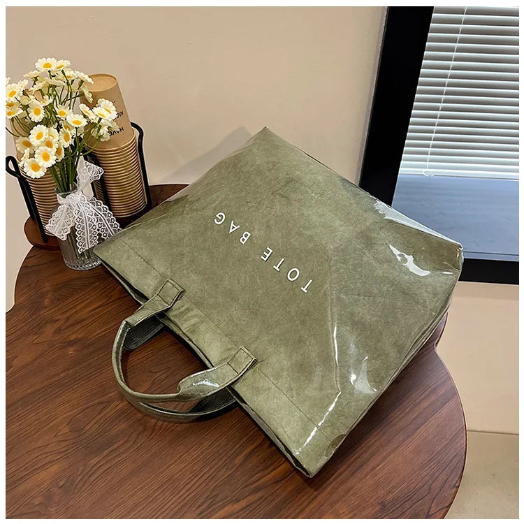 A green transparent waterproof PVC tote bag resting on a wooden table next to a vase of yellow flowers.