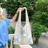 A woman holds a white knitted tote bag with blue embroidery outdoors against a backdrop of green foliage.
