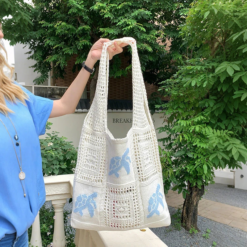 A woman holds a white knitted tote bag with blue embroidery outdoors against a backdrop of green foliage.