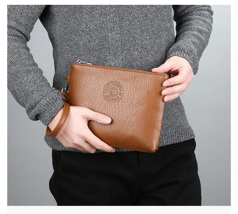 A person holds a brown PU leather men's clutch wallet against a gray sweater and black pants.