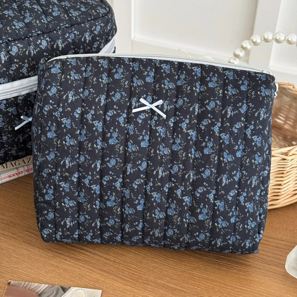 A dark blue floral patterned quilted cosmetic bag with a silver zipper and cross detail resting on a wooden surface.