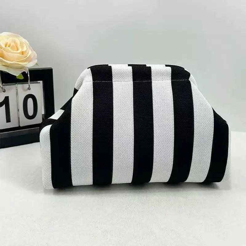 A black and white striped knit cloud makeup bag resting on a white surface next to a yellow rose.