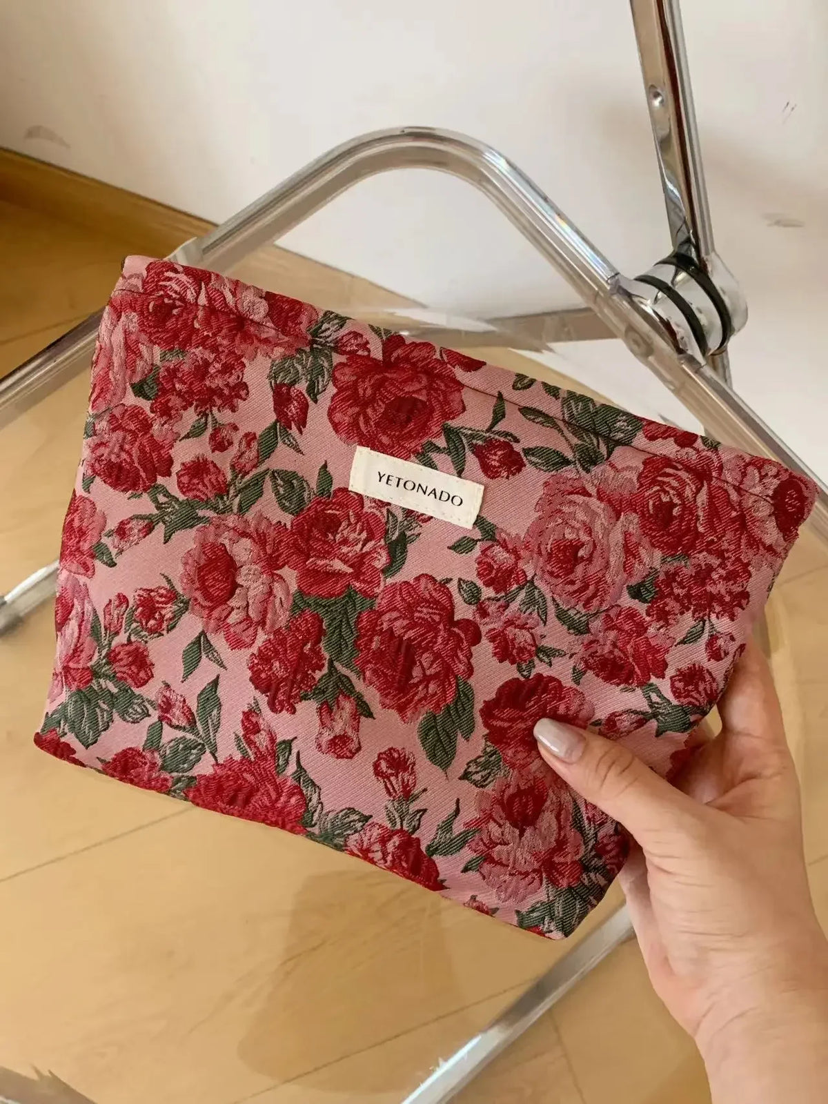 A red floral patterned cosmetic pouch is held in a hand, resting on a glass or metal frame.
