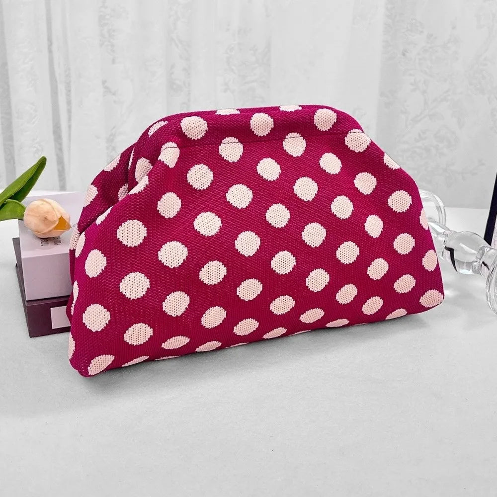 A magenta polka dot clutch bag is displayed on a light surface next to some clear glass.