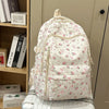 Mochila blanca con estampado floral y múltiples bolsillos sobre una superficie clara junto a una estantería.