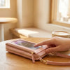 Eine Hand hält eine rosa Leder-Crossbody-Handytasche mit goldener Kette auf einem Holztable.