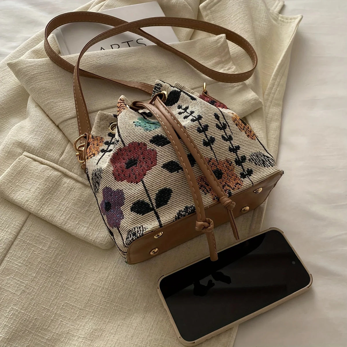 A beige floral drawstring bucket bag with brown leather accents resting on a cream-colored jacket and a smartphone.