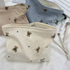 A beige corduroy clutch bag with small brown bear embroidery is shown next to a blue patterned bag and makeup brushes.
