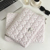A quilted pink laptop sleeve with a zipper and small floral embroidery resting on a white table next to a silver perfume bott