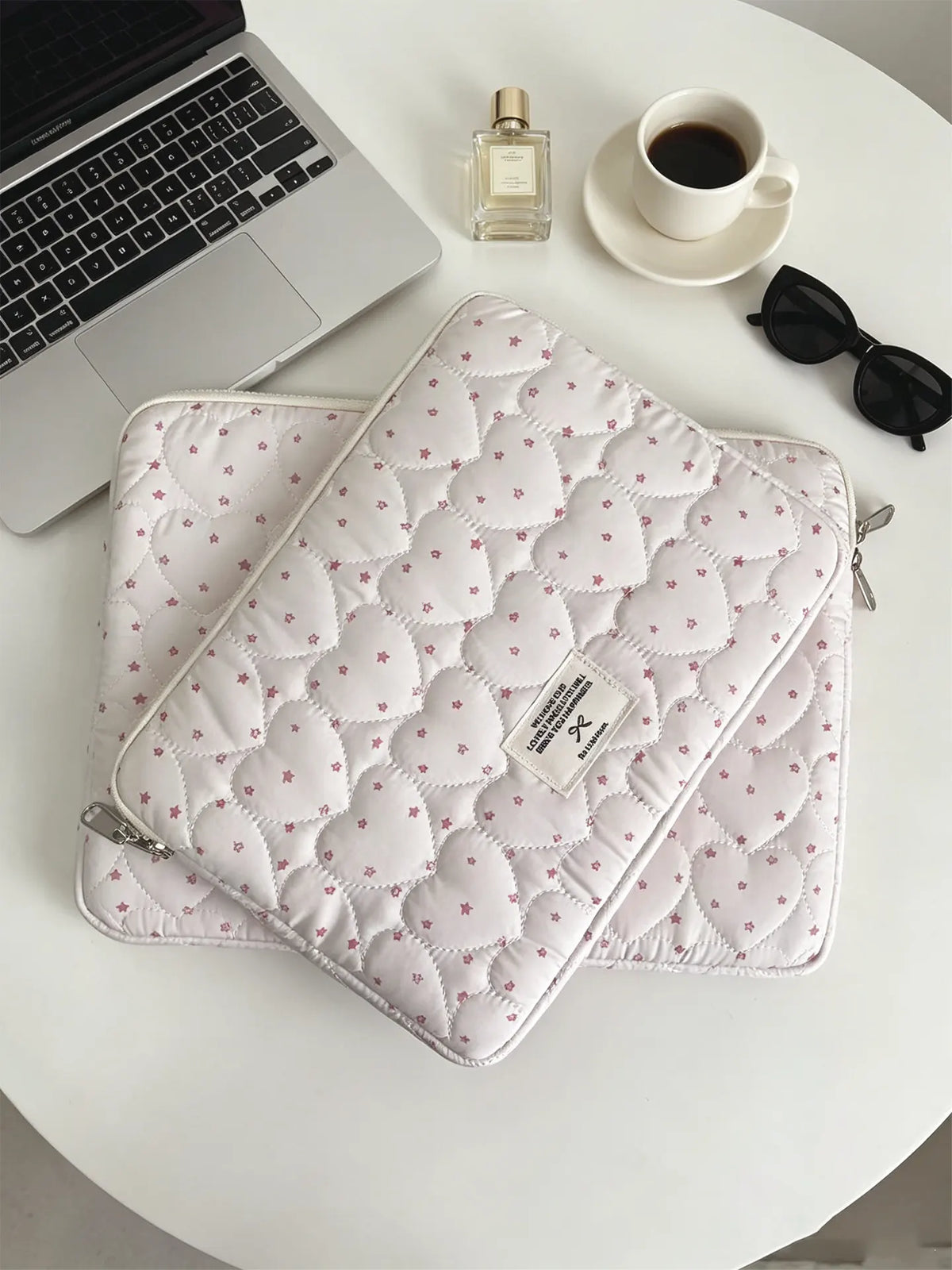 A quilted pink laptop sleeve with a zipper and small floral embroidery resting on a white table next to a silver perfume bott