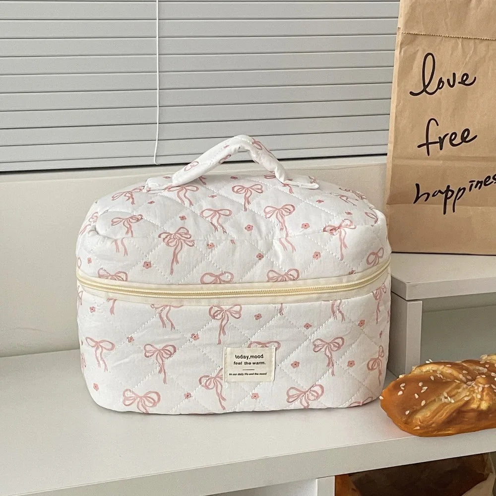 A white quilted cosmetic bag with pink embroidery rests on a shelf next to a piece of bread.