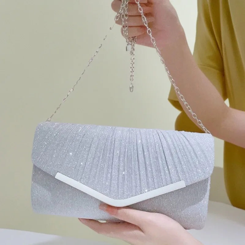A hand holds a silver glitter pleated crossbody clutch bag with a detachable chain against a light background.