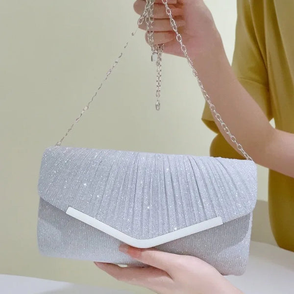 A hand holds a silver glitter pleated crossbody clutch bag with a detachable chain against a light background.
