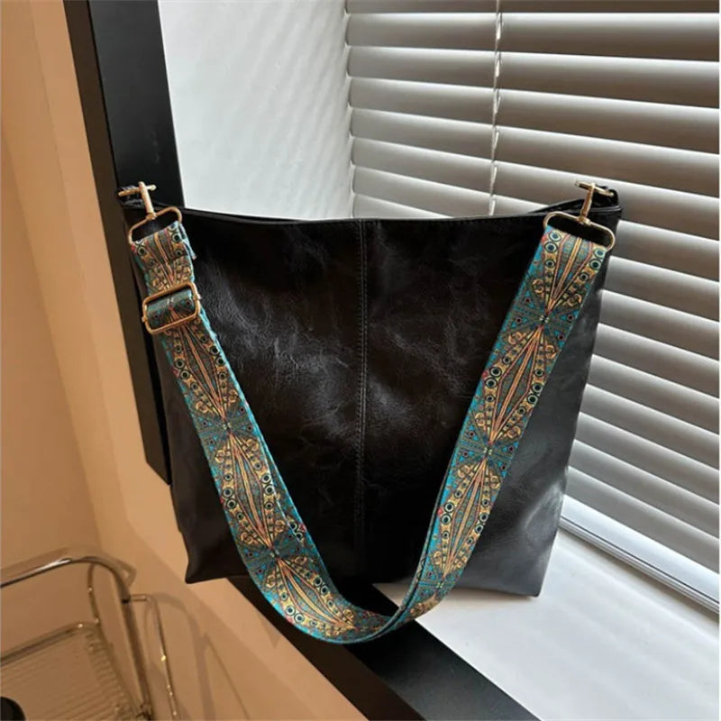 A black shoulder bag with a wide, embroidered strap featuring teal and gold patterns, displayed against a window blind.