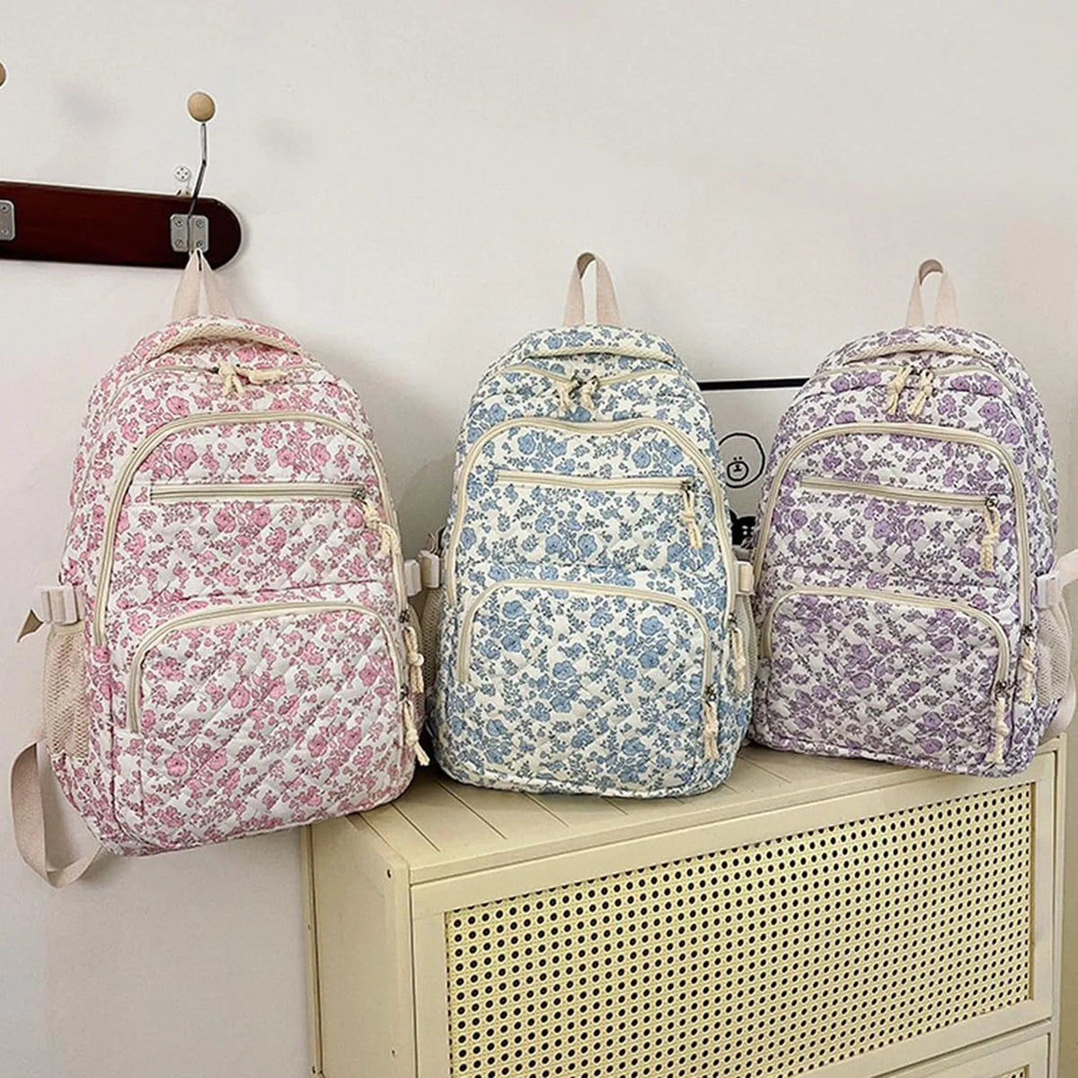Three floral quilted backpacks in pink, blue, and purple are displayed on a shelf.