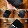 A black minimalist full-grain leather passport holder and card wallet is shown being held on a wooden surface.