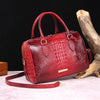 A red crocodile pattern luxury handbag with a leather strap resting on a light surface next to a piece of driftwood.