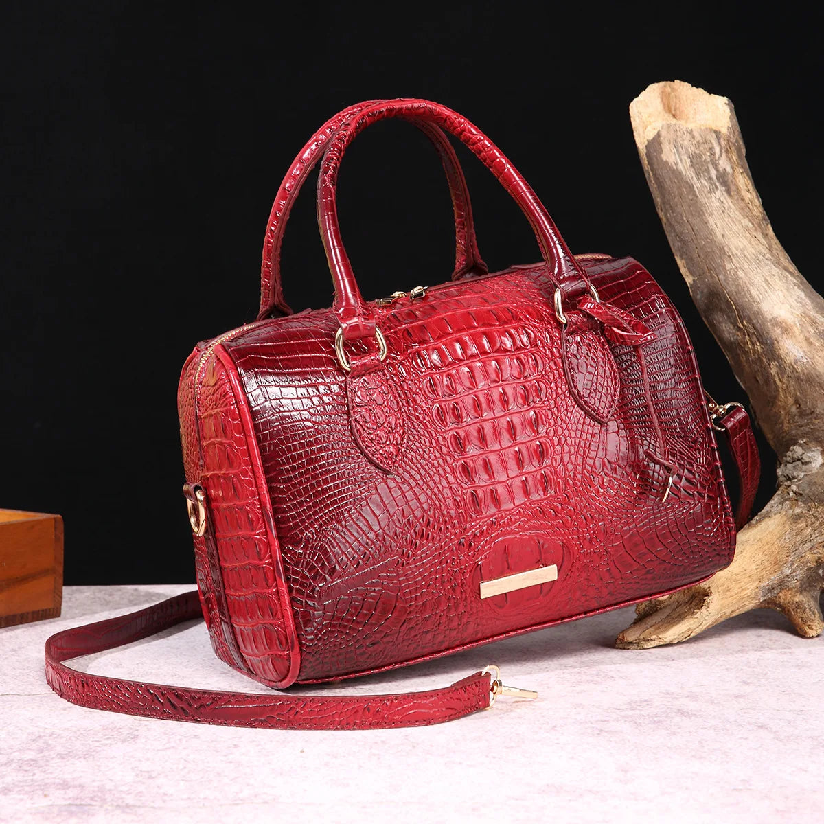 A red crocodile pattern luxury handbag with a leather strap resting on a light surface next to a piece of driftwood.