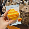 A hand holds a yellow silicone protective case for wireless earbuds against a blurred background of brown silicone products.