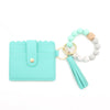 A teal silicone bead bracelet is displayed next to a light wood and white bead bracelet and a leather card holder.