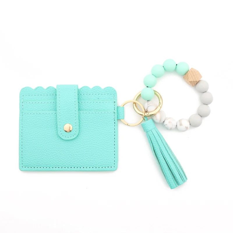 A teal silicone bead bracelet is displayed next to a light wood and white bead bracelet and a leather card holder.