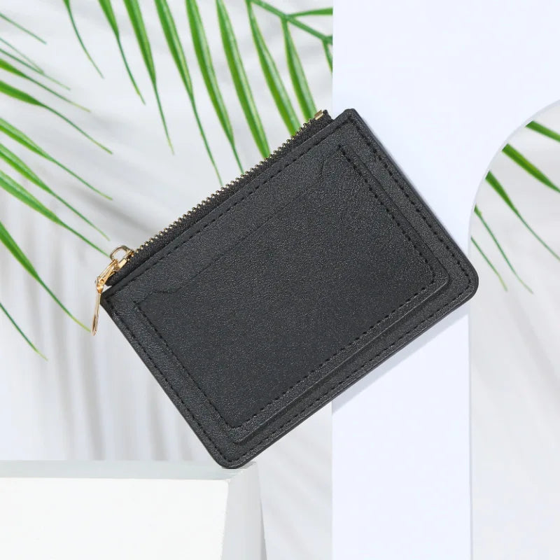 A black PU leather mini card holder wallet with a zipper closure is shown against a white background with green leaves.