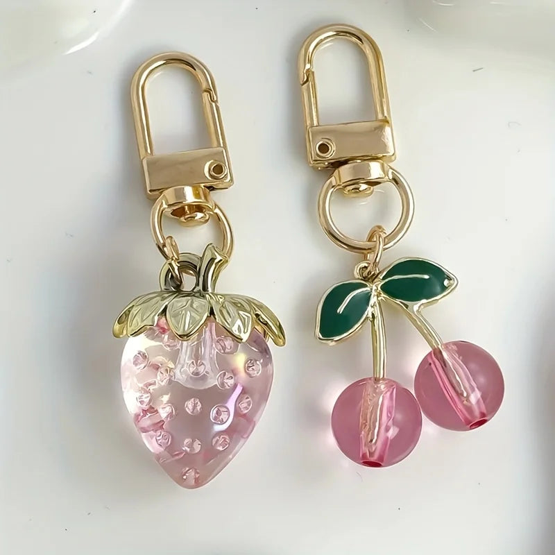 A set of gold-toned fruit keychains featuring a pink strawberry charm and a pink cherry charm.