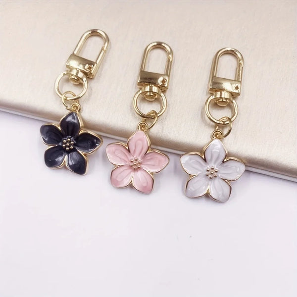 Three gold-toned resin flower keychains in black, pink, and white are displayed on a neutral background.