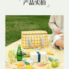 A yellow and orange insulated picnic tote is shown on a checkered cloth with various food items like bread, fruit, and drinks