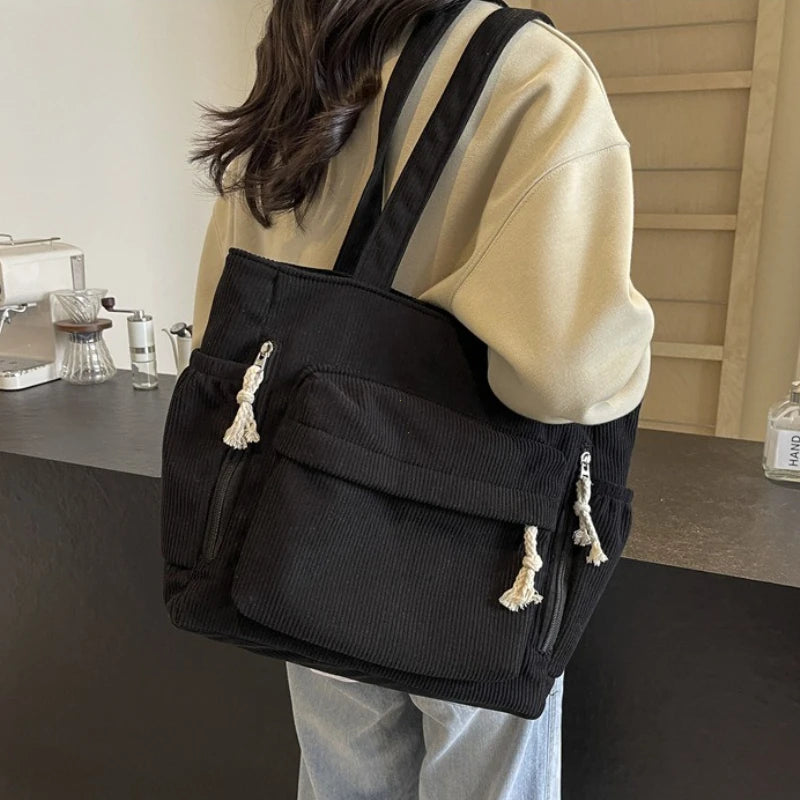 A black corduroy shoulder bag with zipper and tassels is shown carried on a person's shoulder.
