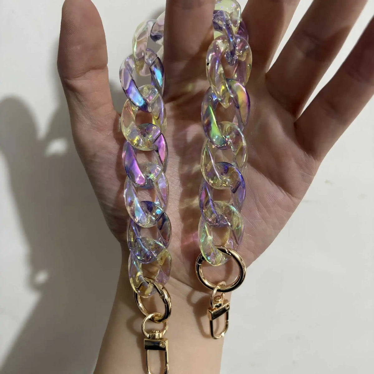 A hand holds a long, iridescent resin chain bag strap with gold-toned charms.