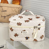 A white cosmetic makeup bag with a quilted pattern and brown bear appliqués sits on a white shelf.