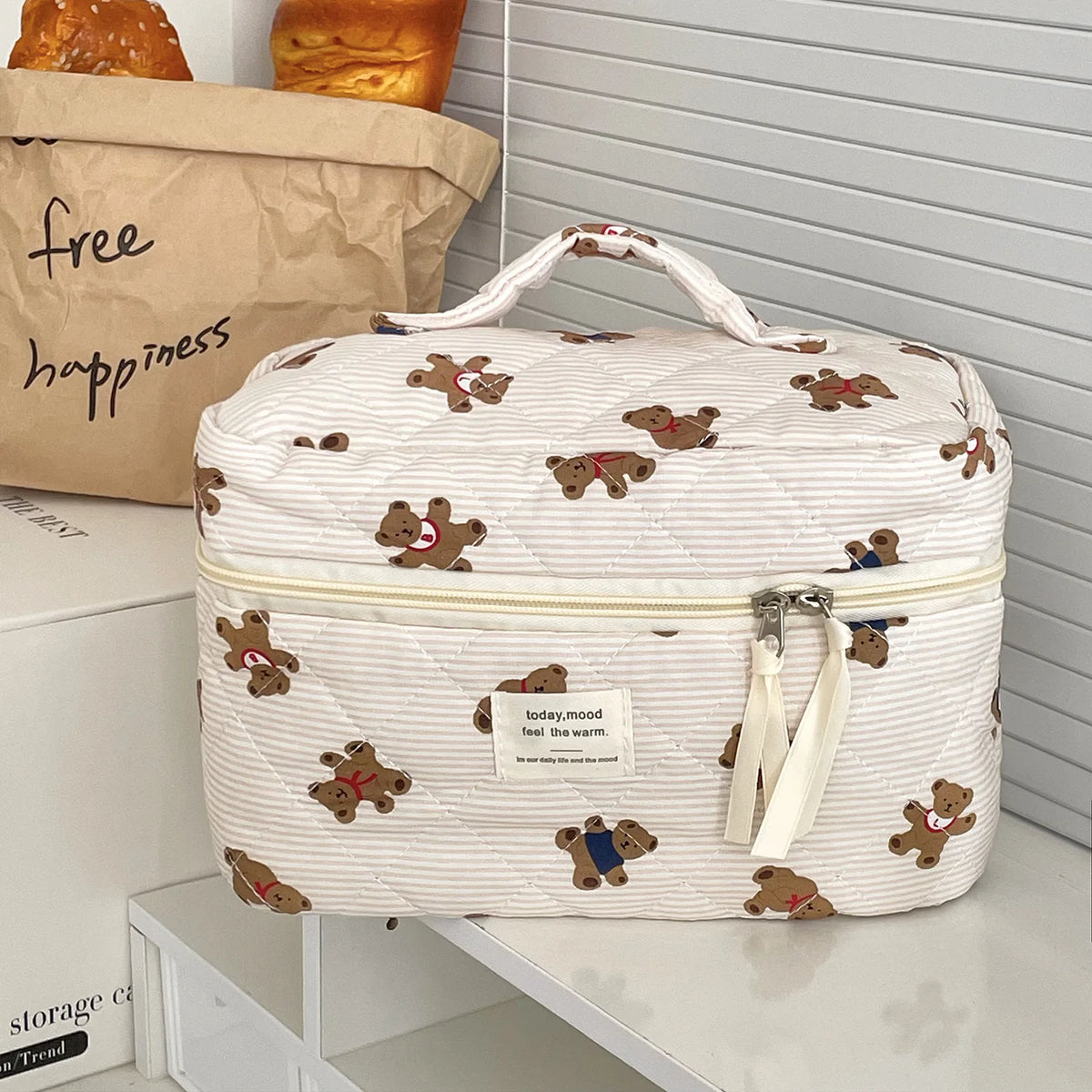 A white cosmetic makeup bag with a quilted pattern and brown bear appliqués sits on a white shelf.