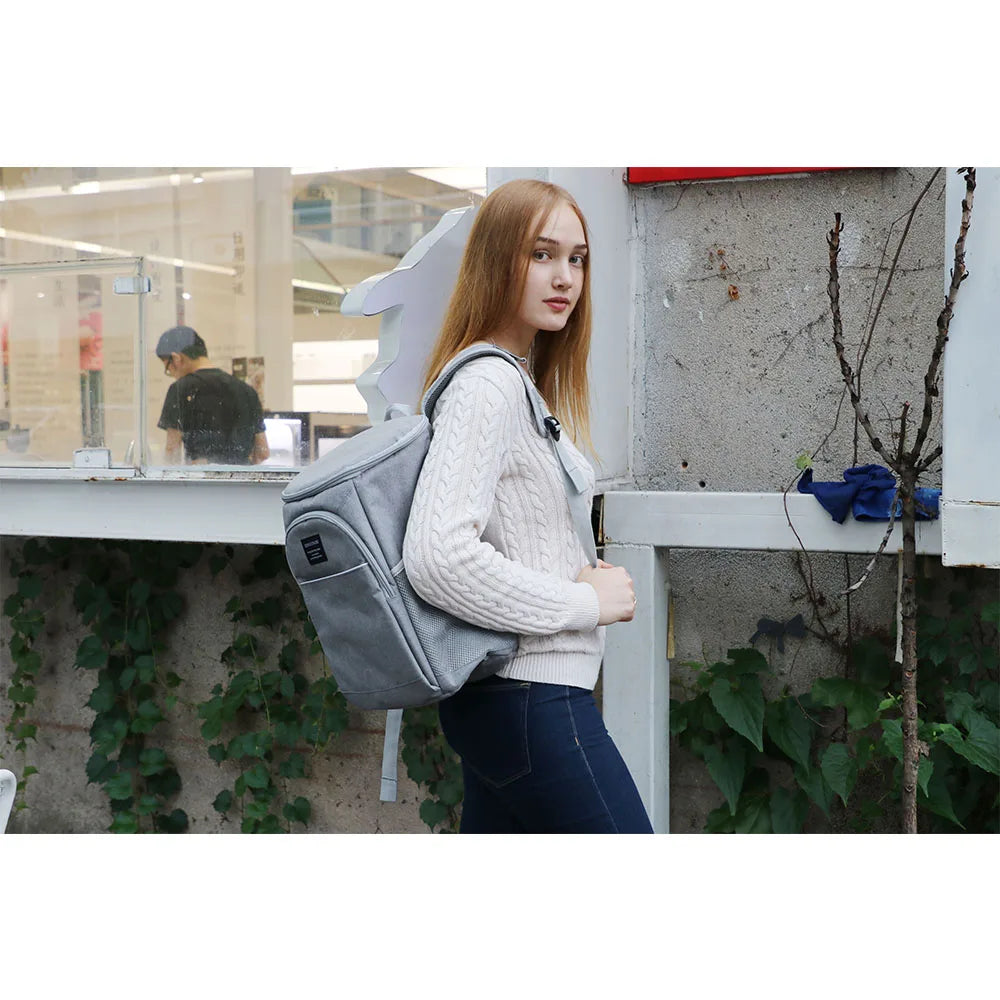 A young woman with long blonde hair is standing outdoors holding an Insulated Thermal Cooler Backpack against a light-colored