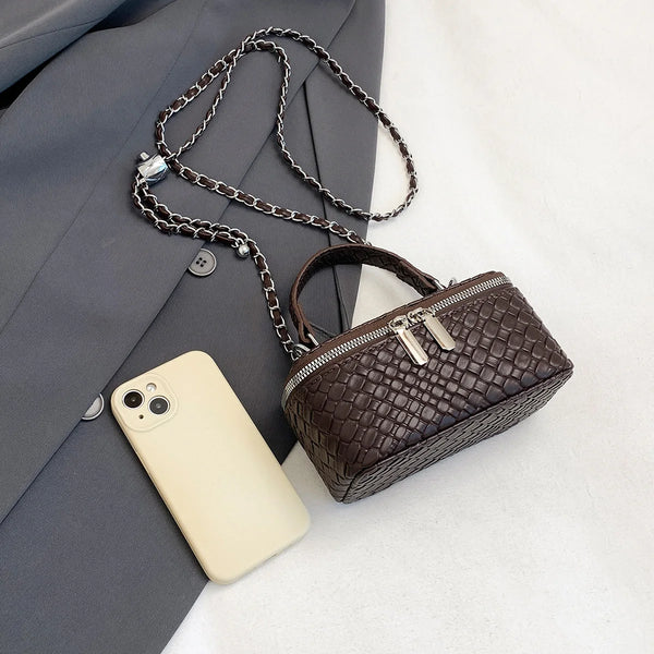 A dark brown woven crossbody bag with a silver chain strap resting next to a light-colored smartphone and a gray blazer.
