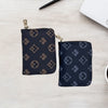 A dark blue patterned zippered card holder wallet resting on a light wooden surface next to a pen and a white notebook.
