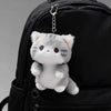 A grey soft cat plush pendant is hanging from a zipper pull on a black backpack.