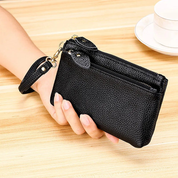 A hand holds a black genuine leather wristlet clutch wallet with a zipper closure on a wooden surface.
