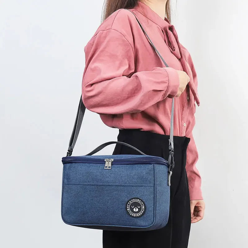 A woman holds a navy blue insulated portable lunch bag with a shoulder strap against a white background.