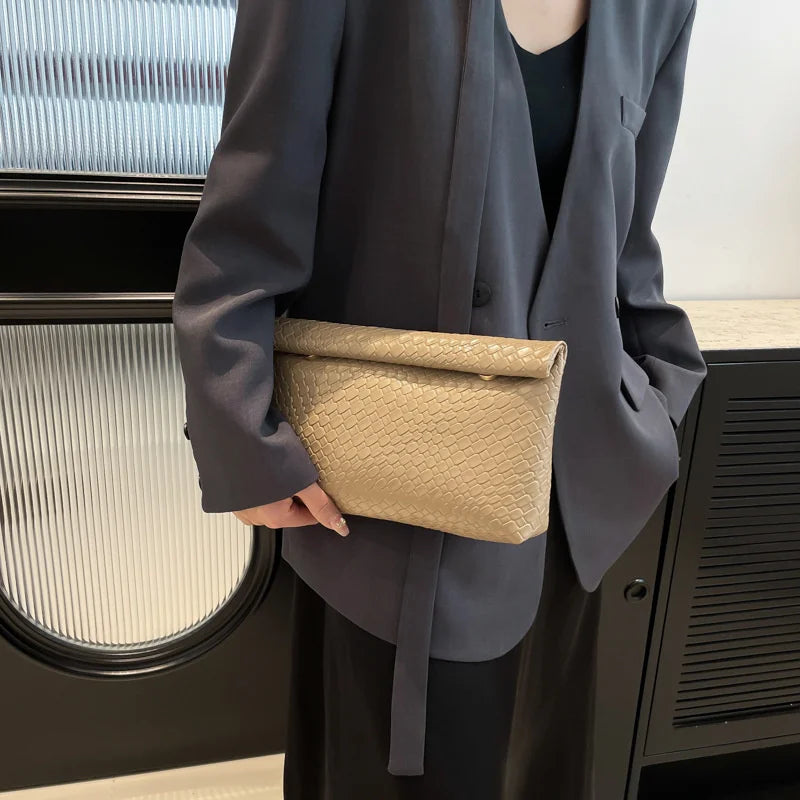 Una persona sostiene un elegante bolso de mano tejido color beige claro contra un blazer gris oscuro.