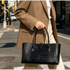 A woman holds a black crocodile embossed leather tote bag in a light brown blazer while walking on a city street.