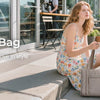 A woman wearing a light floral dress sits on a sidewalk, holding a beige quilted tote bag.